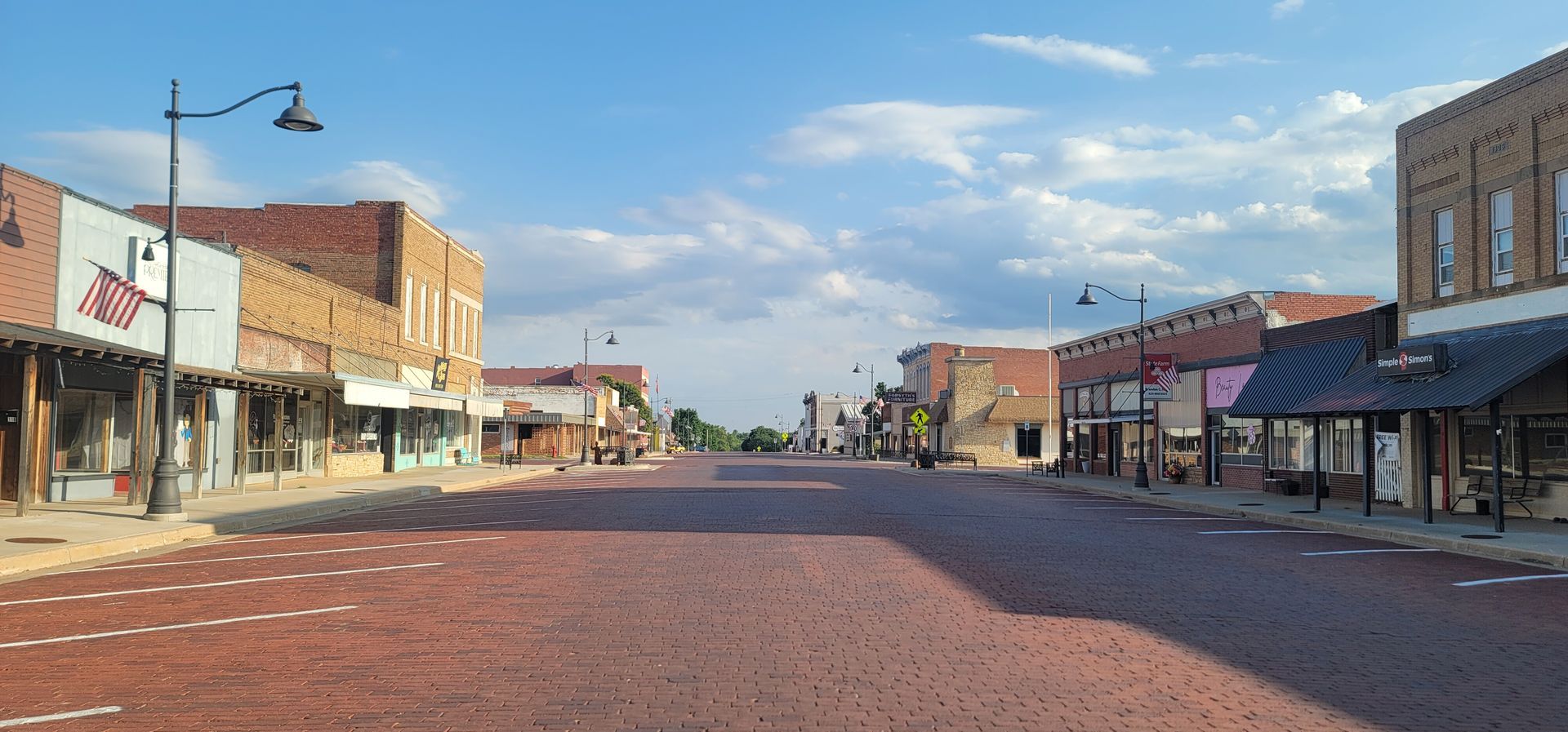 Quiet small-town main street with brick road, storefronts, and streetlamps under a blue sky.