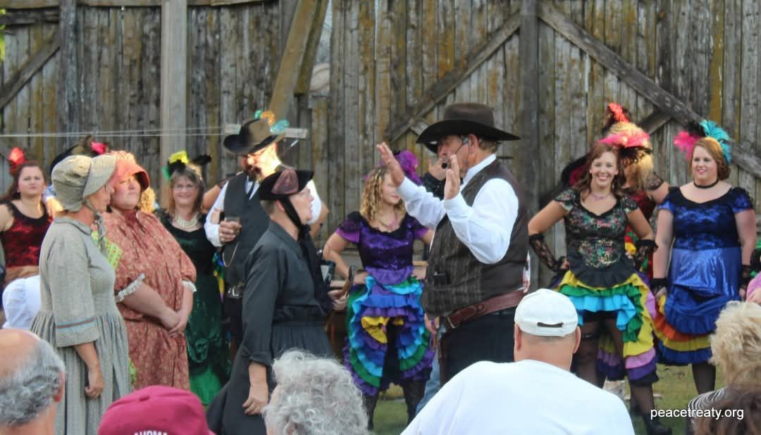 Performers in cowboy hats and colorful costumes singing on an outdoor stage before a crowd