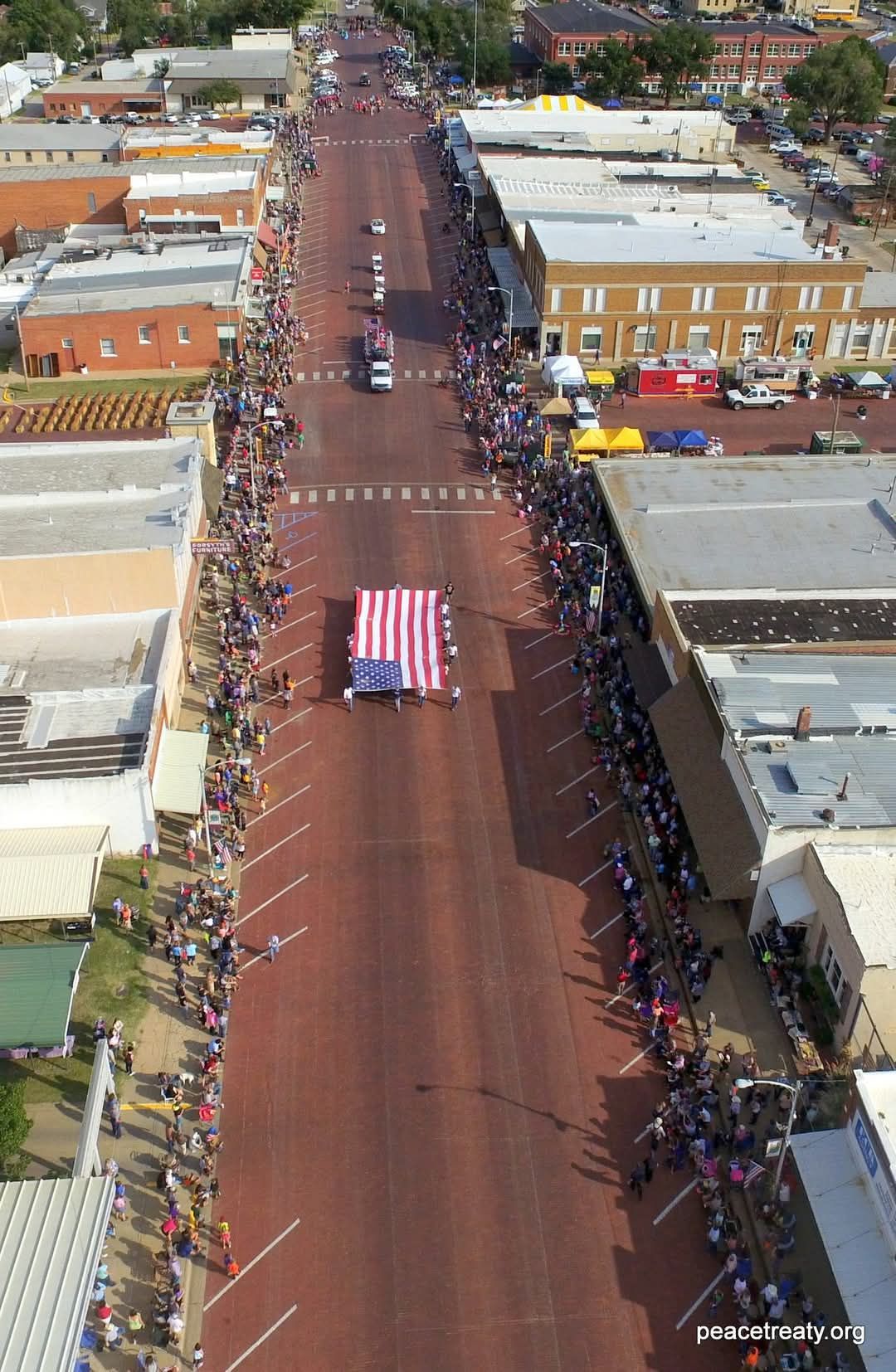 Aerial view of a long street lined with crowds and buildings, with a large U.S. flag banner in the center.
