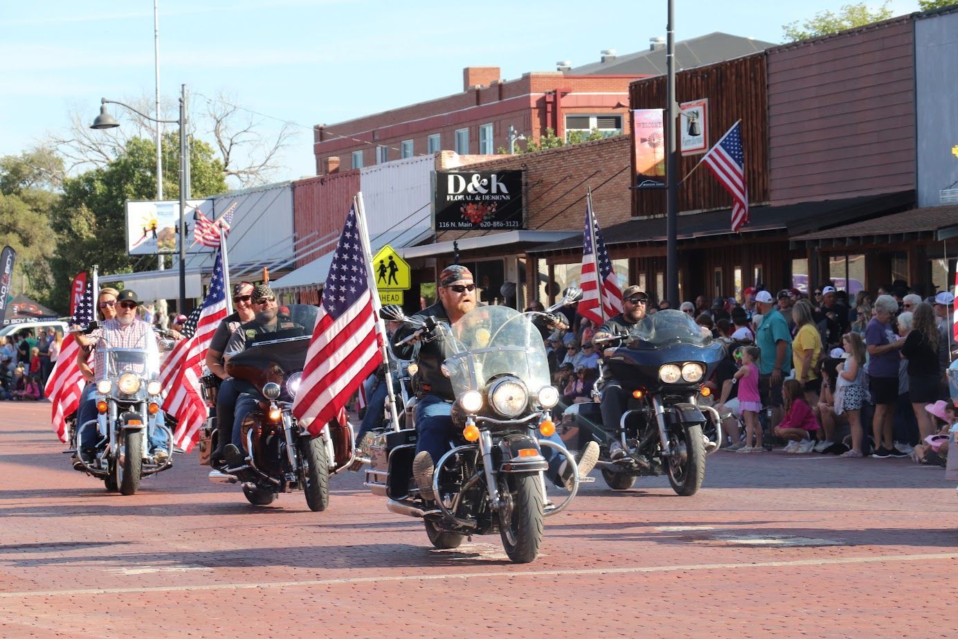 Motorcycle parade with riders carrying American flags down a downtown street during a crowd-lined event