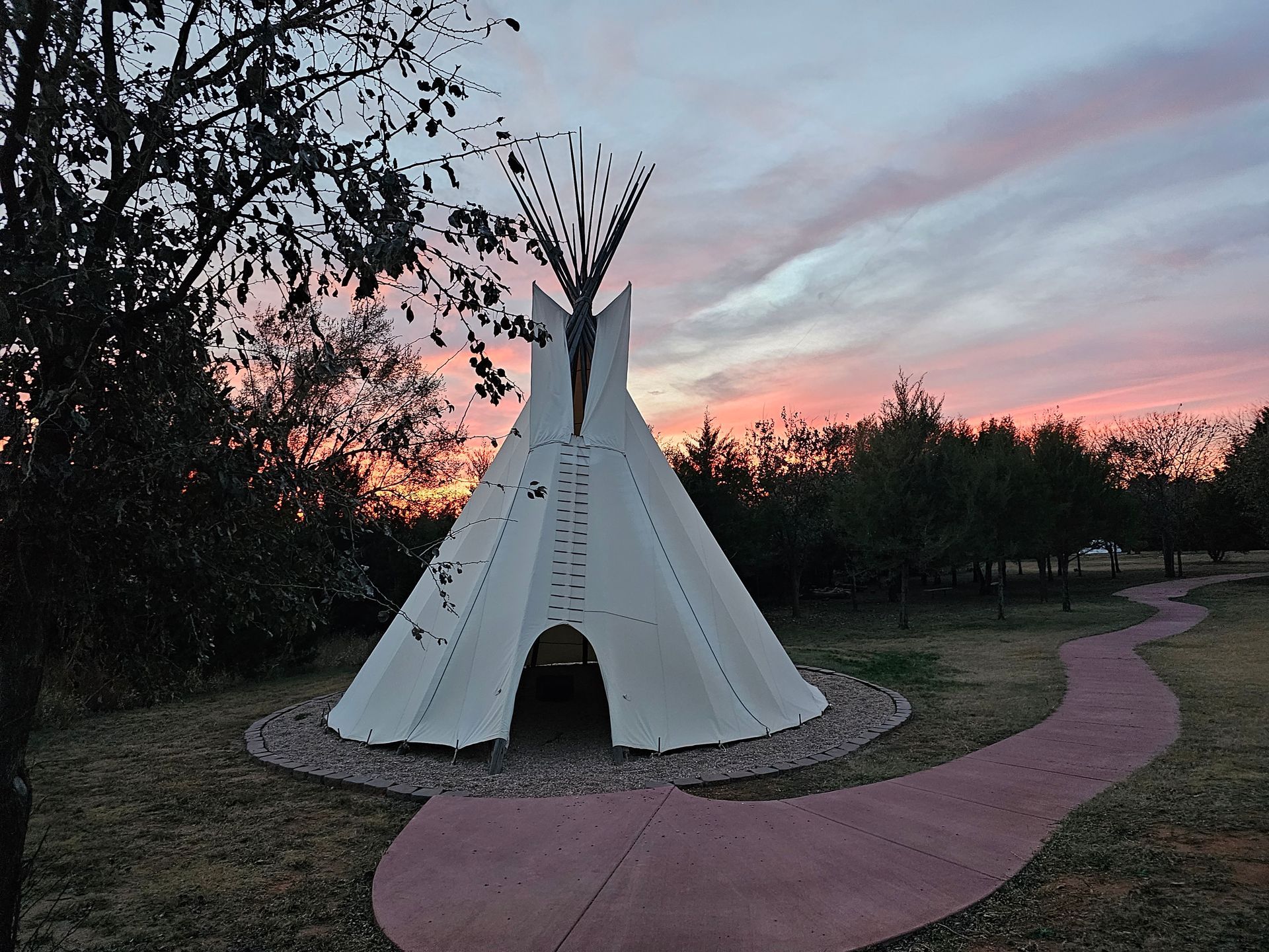 A white teepee-like structure on a pink path at sunset, surrounded by trees and a colorful sky.