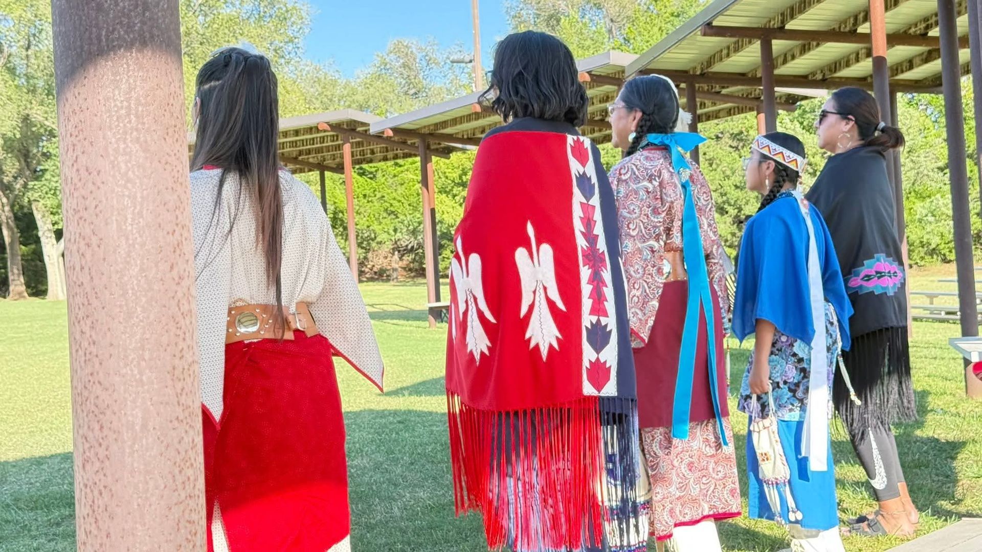 Five people standing outdoors near a pavilion, wearing colorful garments and facing away from the camera