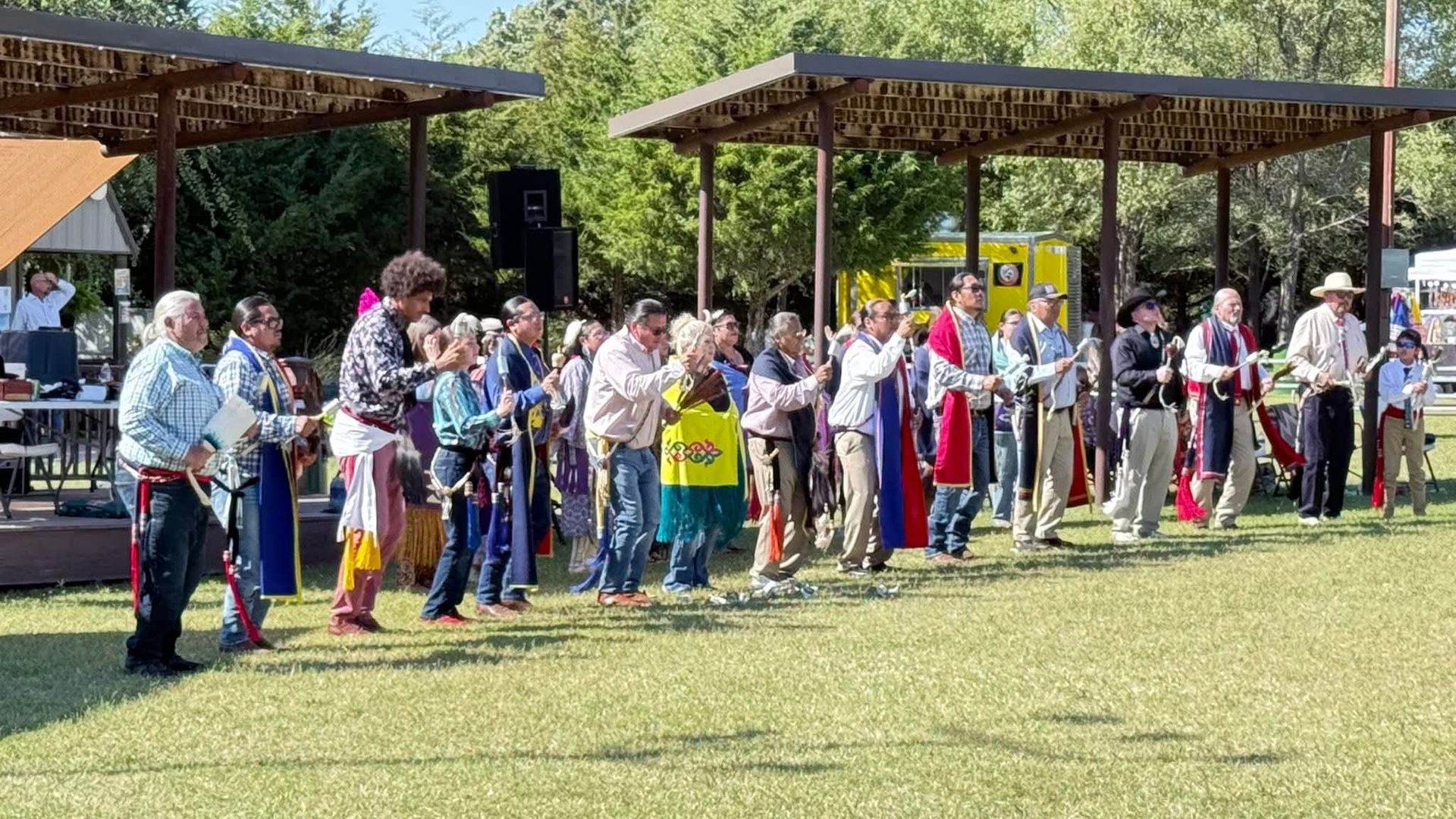 People standing in a line outdoors under picnic shelters, holding colorful fabric strips on a grassy field.