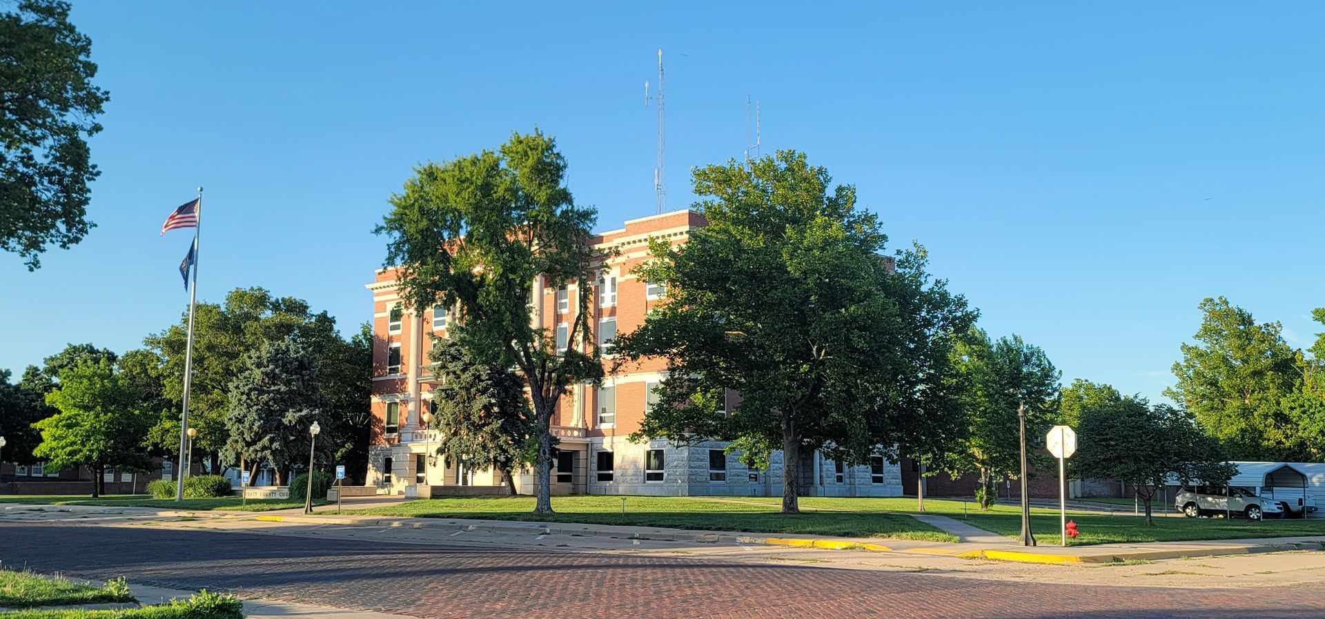 Sunlit civic building with trees, parked cars, and a flag waving under a clear blue sky