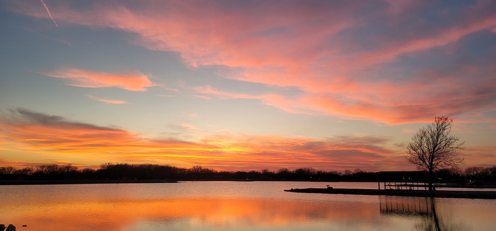 Colorful sunset over a calm lake with pink clouds and a silhouetted dock and tree