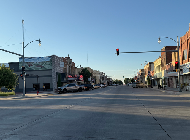 Empty downtown street at a red light, with storefronts and parked cars under a clear blue sky.