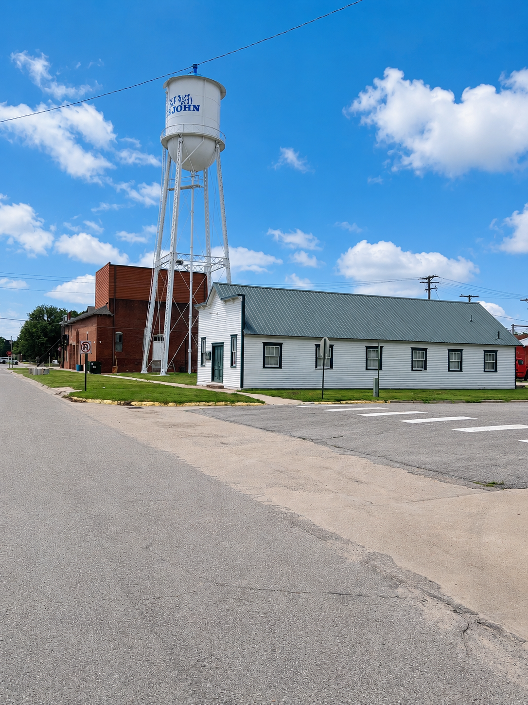 Rural street with a white water tower beside a red brick building and a gray-roofed white building under a blue sky.