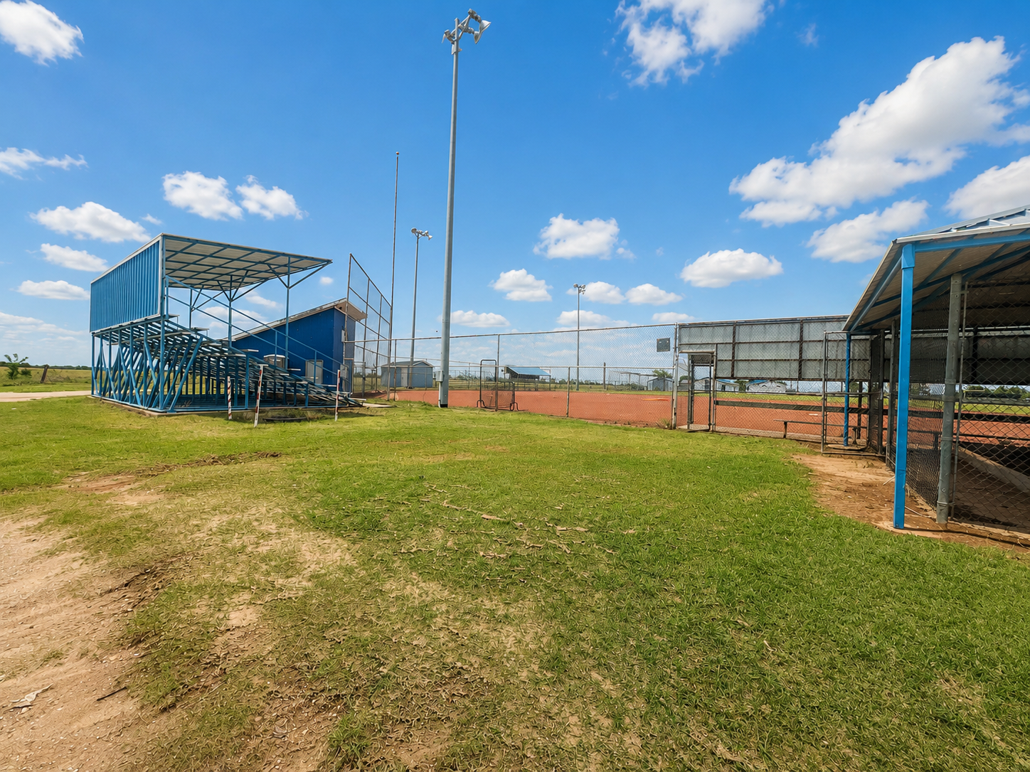 Empty sports field with blue bleachers, fencing, and tall light poles under a sunny sky