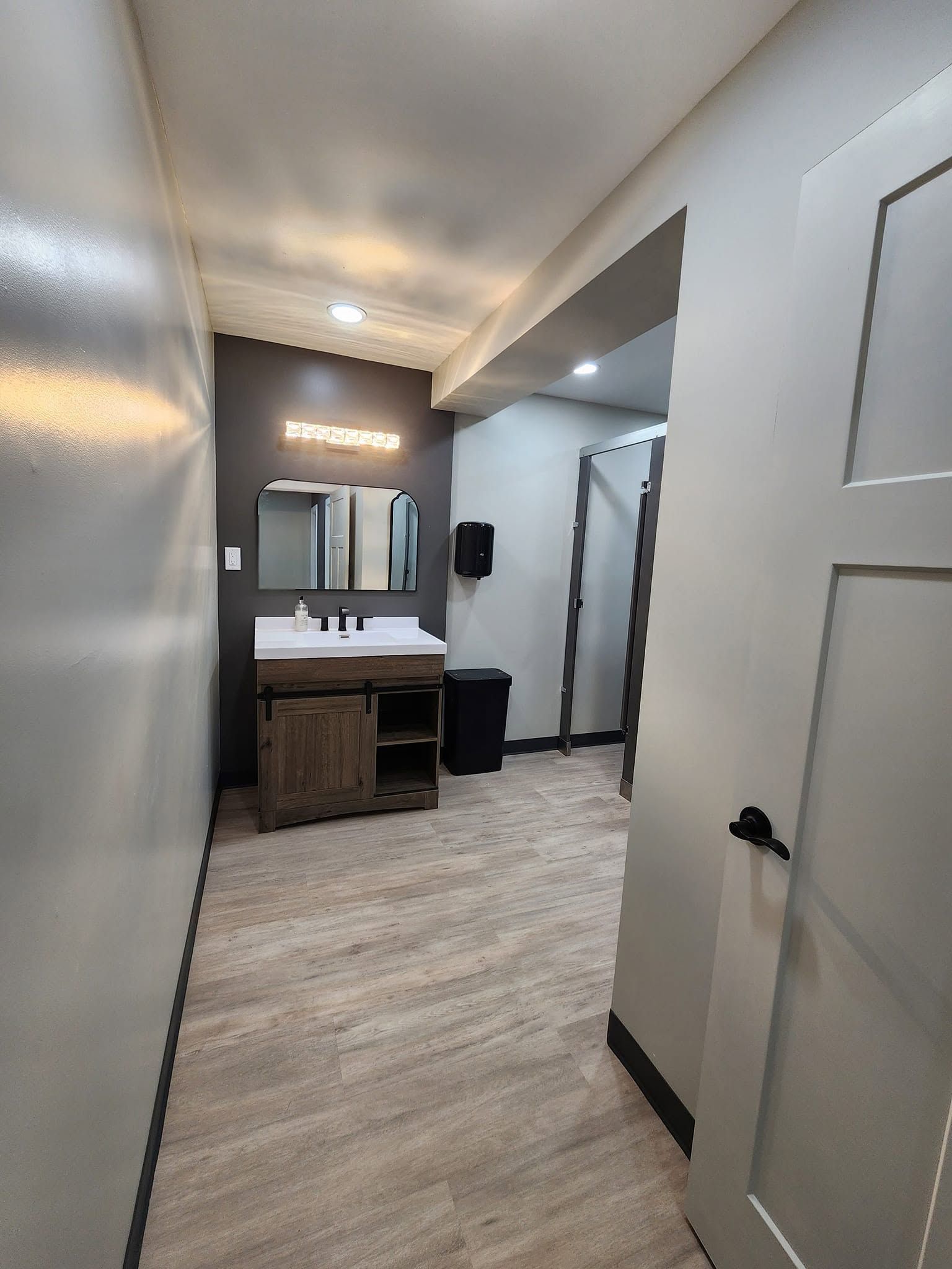 Narrow hallway leading to a bathroom vanity with mirror, lighted ceiling, and open door in a modern interior.