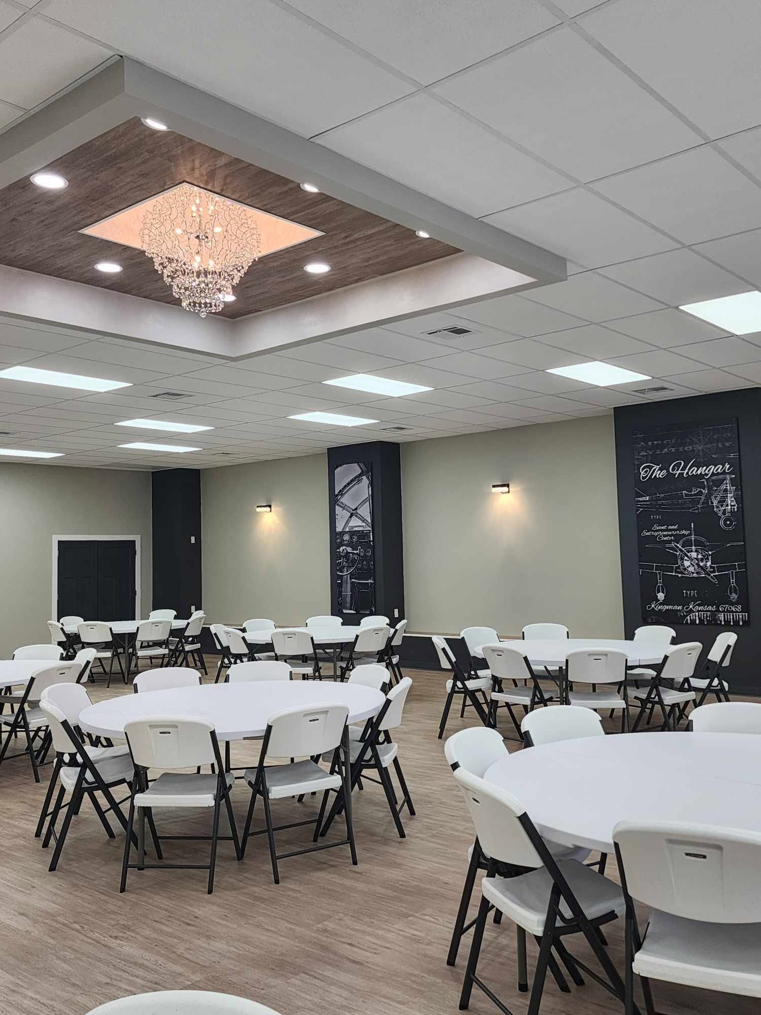 Banquet hall with round tables, folding chairs, and a chandelier under recessed ceiling lights