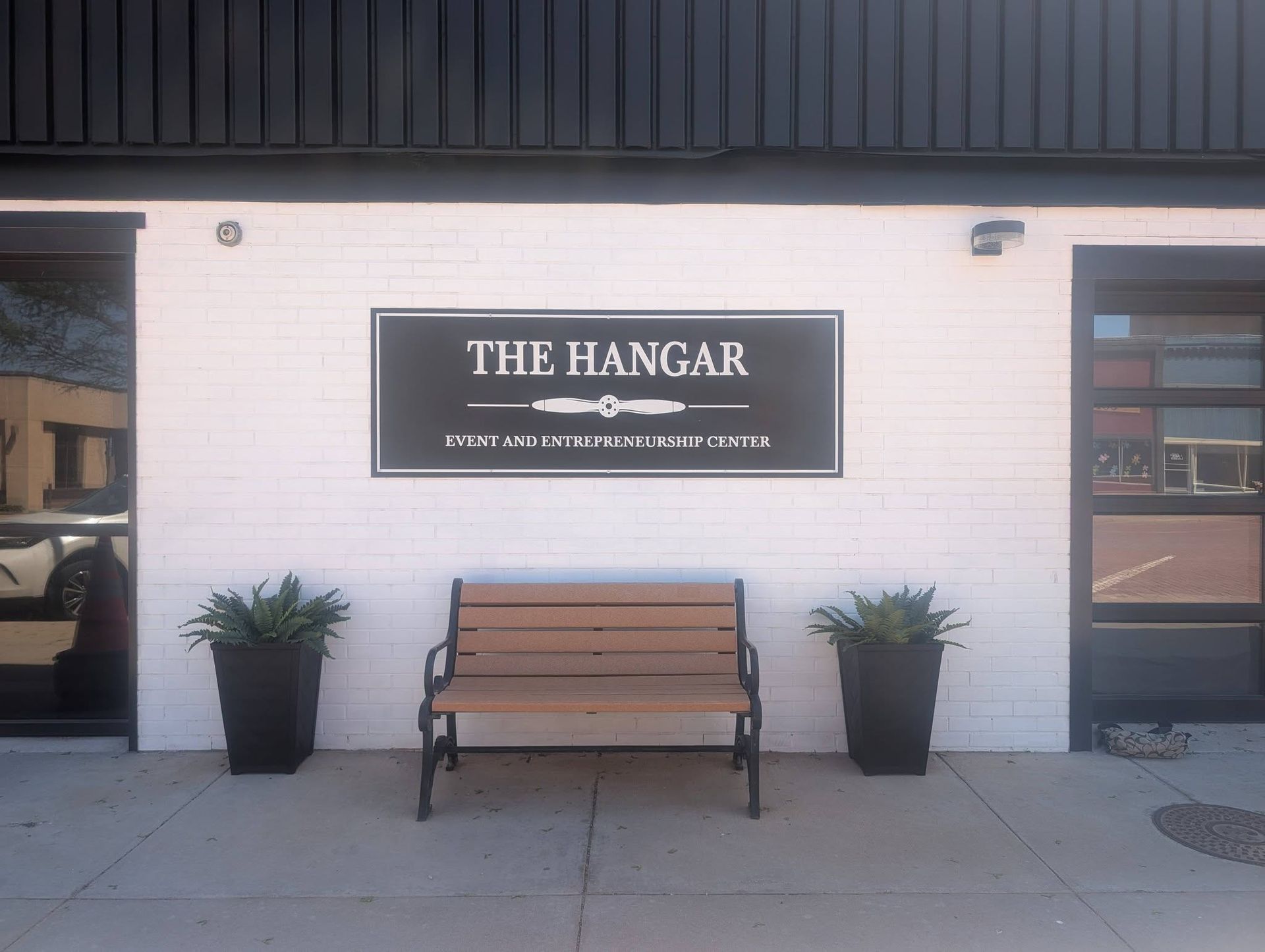 Storefront sign for “The Hangar” above a wooden bench and two potted plants on a white brick wall.