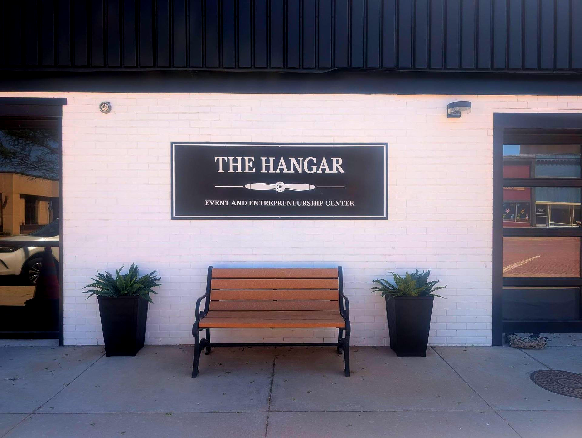 Storefront entrance with “The Hangar” sign, bench, and two potted plants on a white brick wall