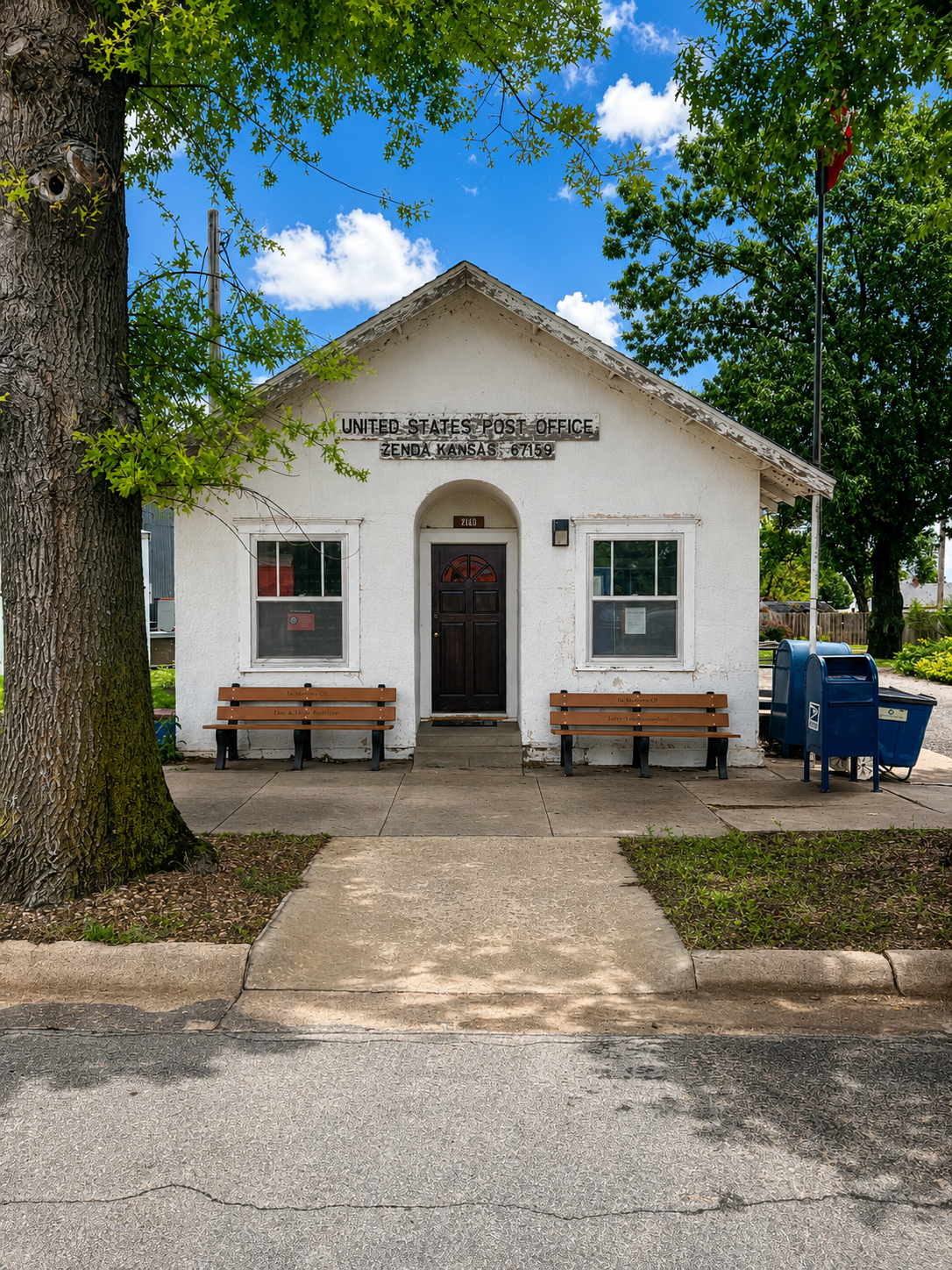 Small white building with bench seating, centered walkway, and blue recycling bins under trees