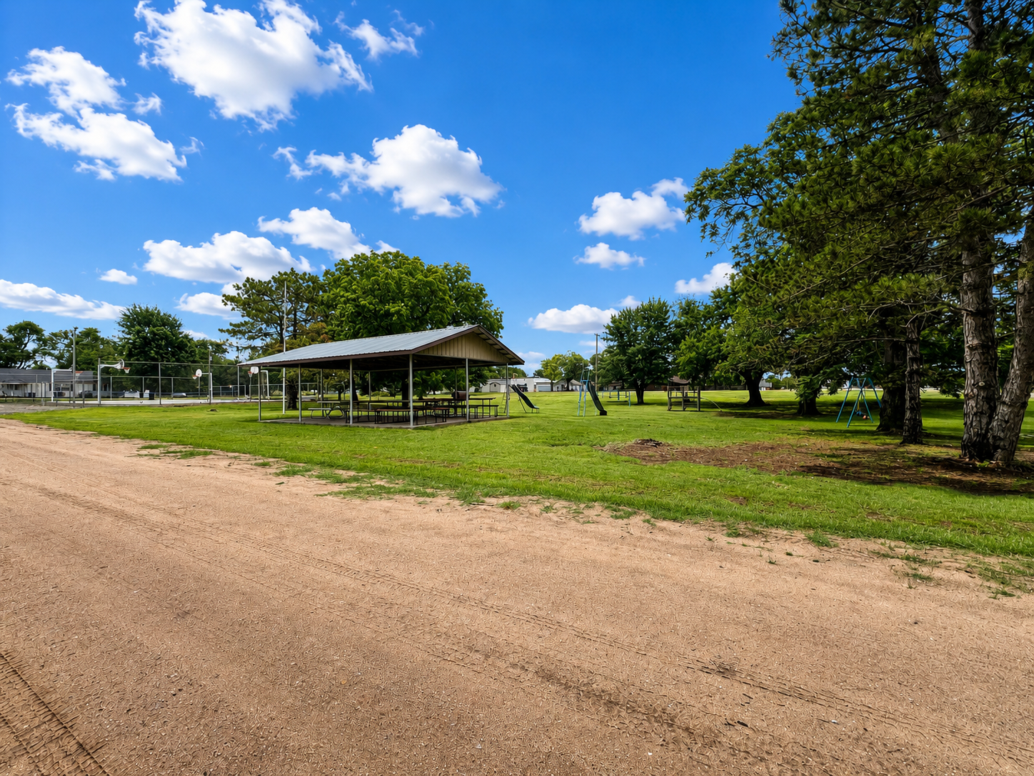 Rural roadside scene with a gravel road, open pavilion, green trees, and a blue sky with clouds