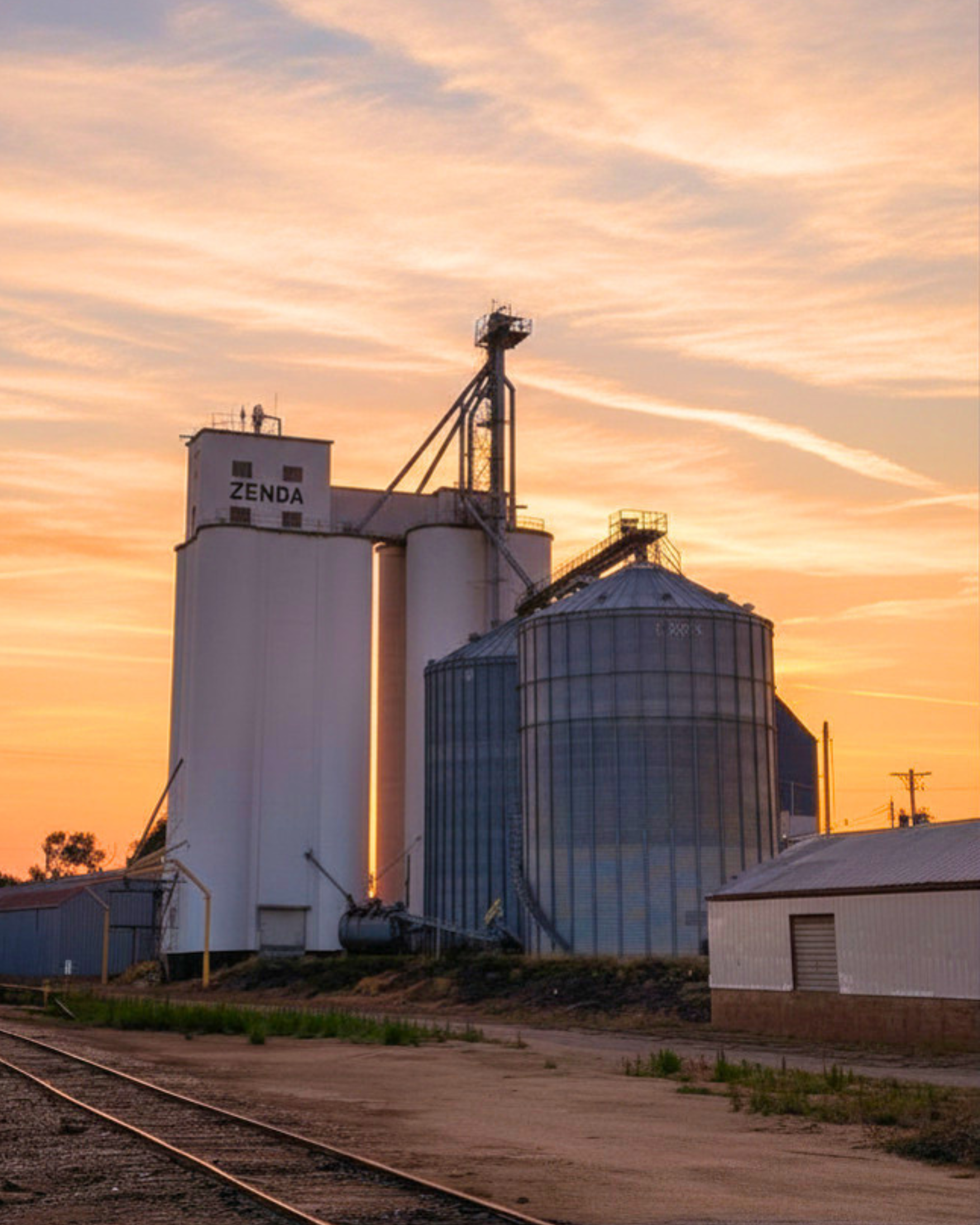 Grain silos and industrial buildings at sunset beside railroad tracks under a pink-orange sky