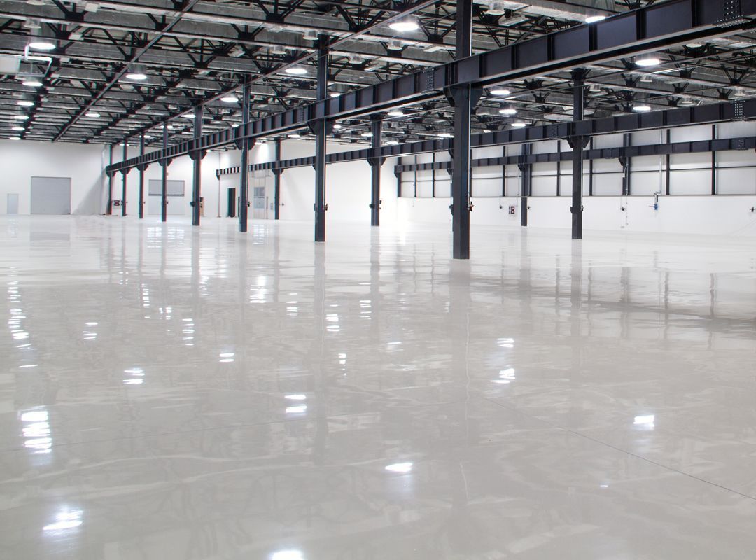 Large, empty warehouse interior with a shiny, light-colored floor, steel beams, and rows of black support columns.