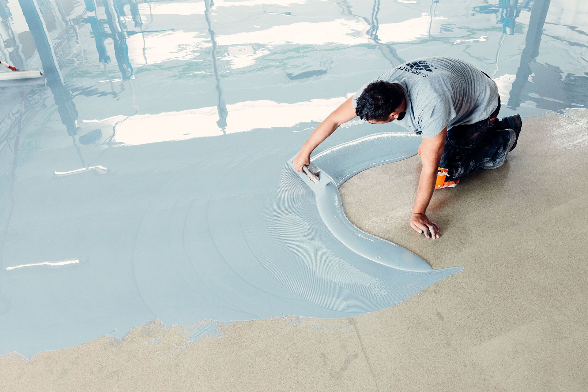 Man applying light blue epoxy coating to a concrete floor with a trowel, in a well-lit industrial setting.