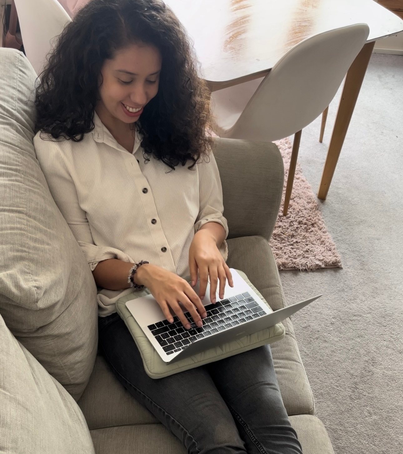 Woman sitting on a bed with a laptop, wrist watch, and phone, working. Neutral tones.