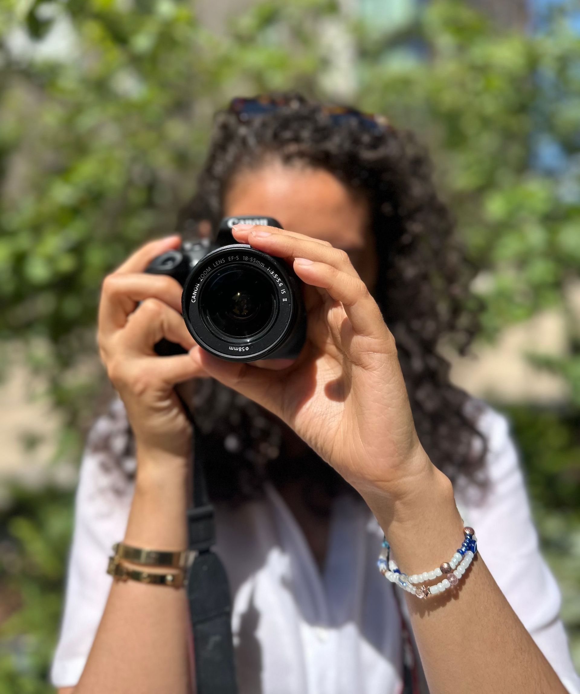 Woman with curly hair taking photo with a camera, outdoors. Wearing bracelets, white shirt, and gold watch.