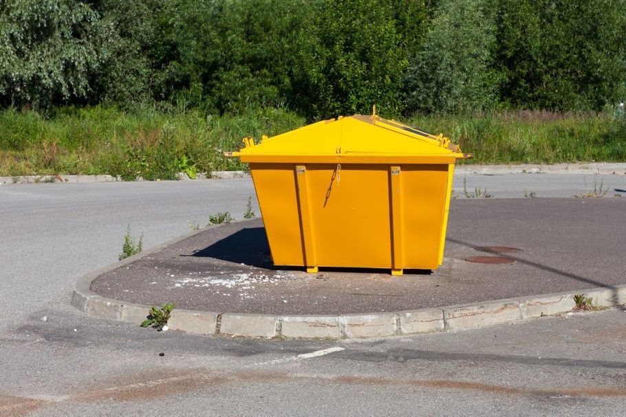 Bright yellow dumpster on a curb island beside a road, with trees in the background