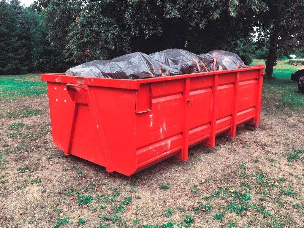 Large red dumpster filled with trash bags on grass near trees