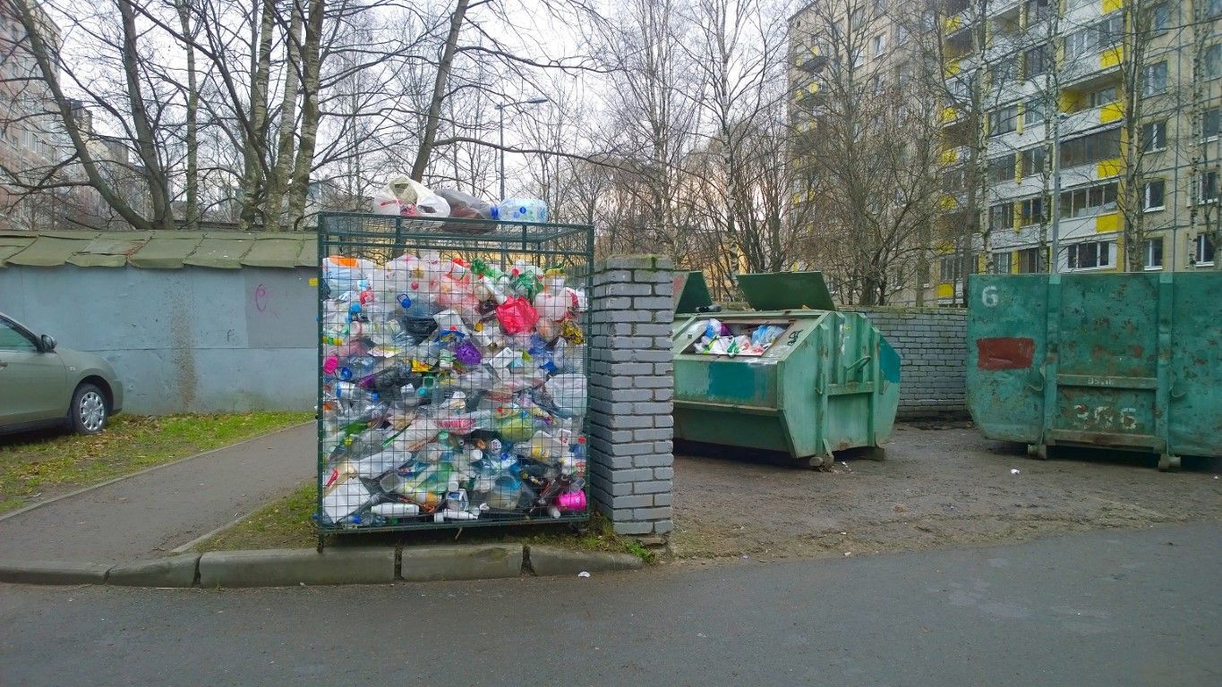 Recycling bins and a colorful paper collection container on a street beside apartment buildings