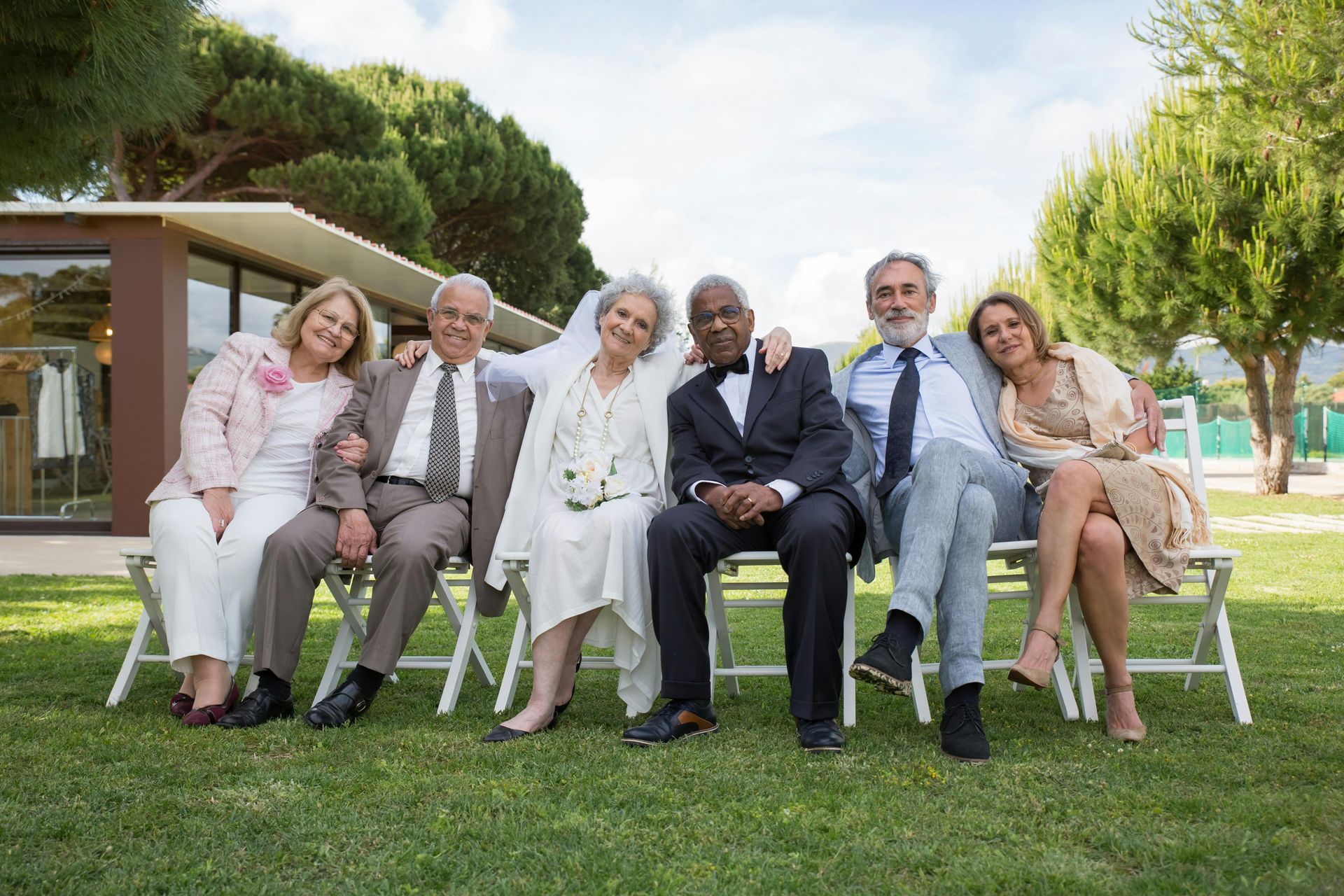 Group of six people seated on chairs outdoors; smiles, green grass, building in background.