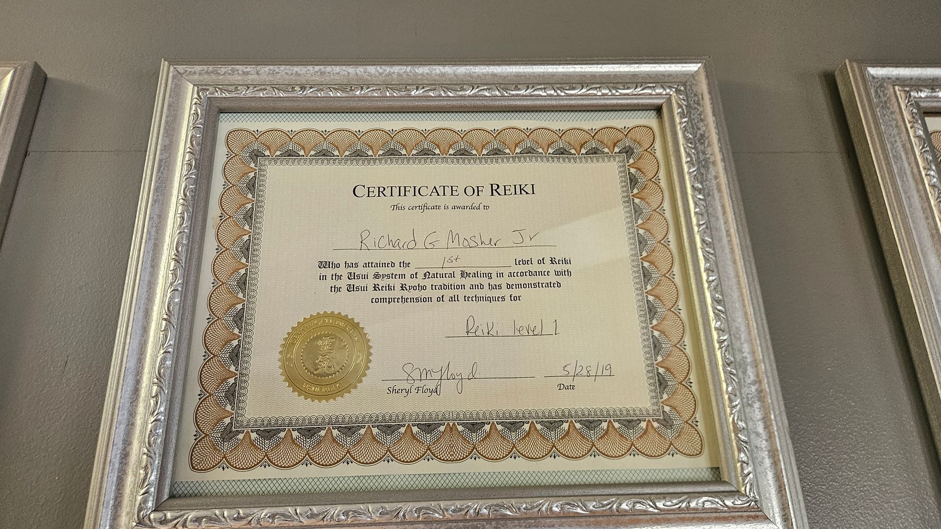Framed certificate with gold seal and ornate border displayed on a gray wall