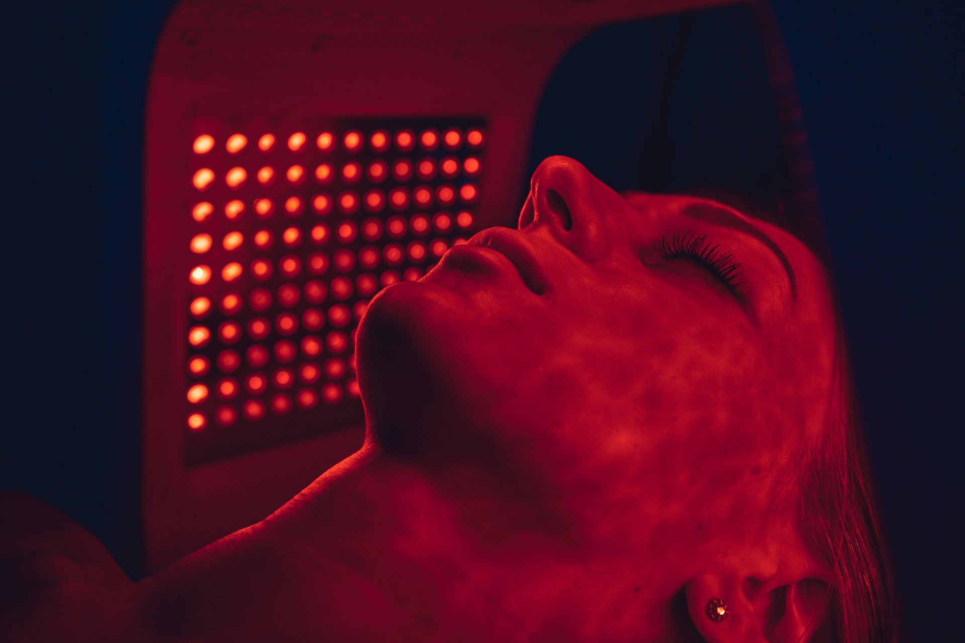 Person lying under red LED light panel, face illuminated in a dark room