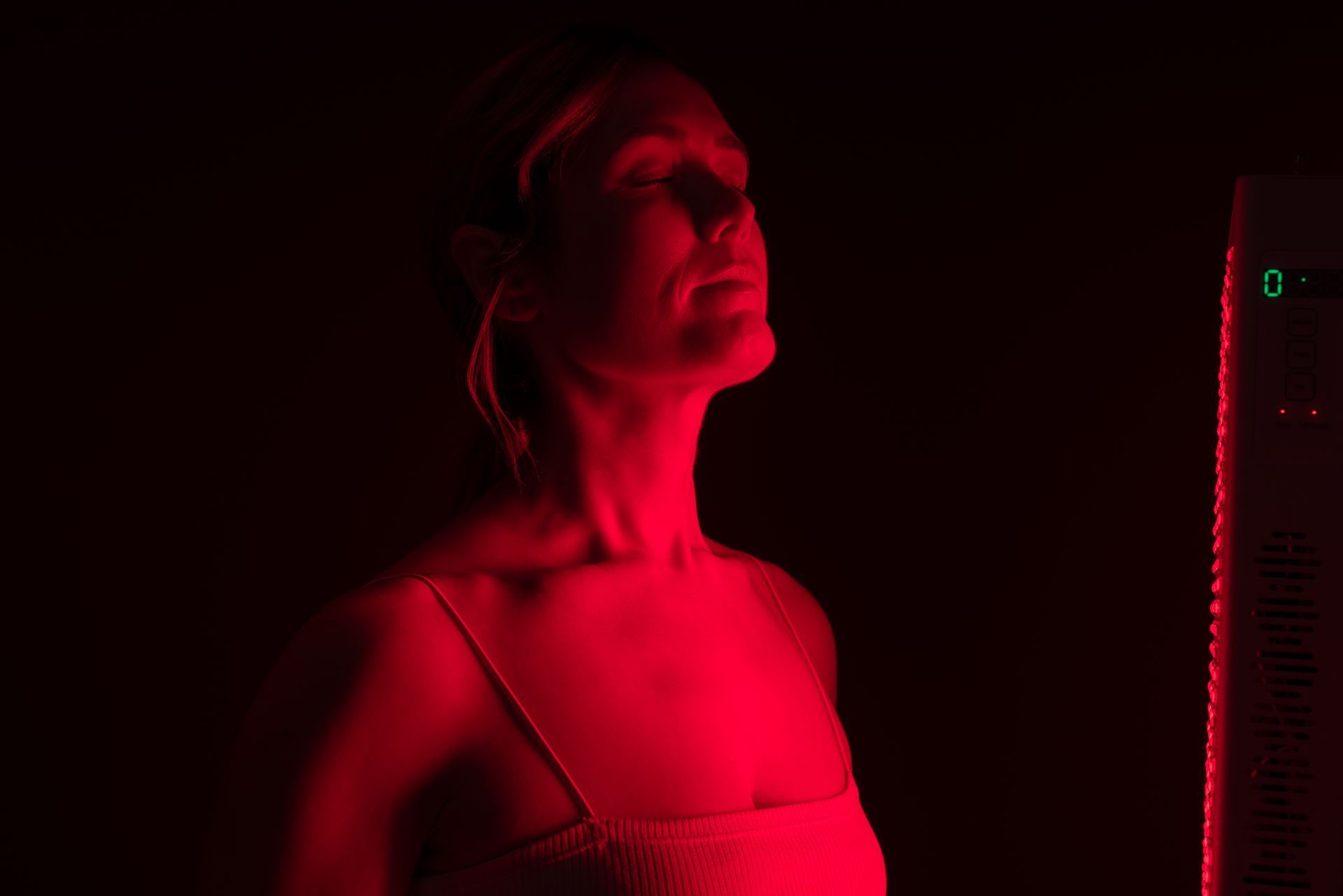 Woman lit by red light looking upward beside a vertical LED panel in a dark room