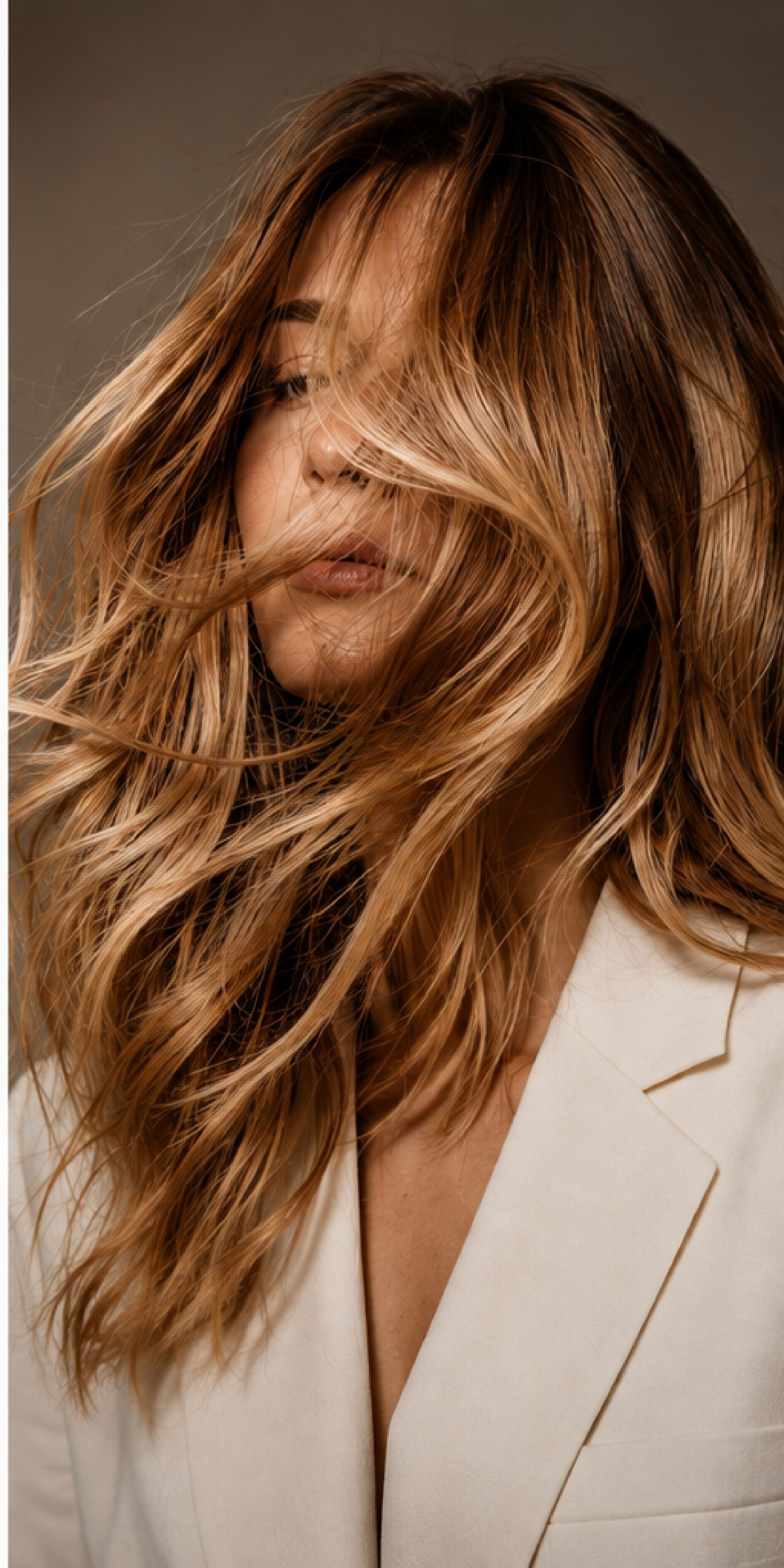 Woman with long wavy brown hair in a cream blazer, partially covering her face against a plain background