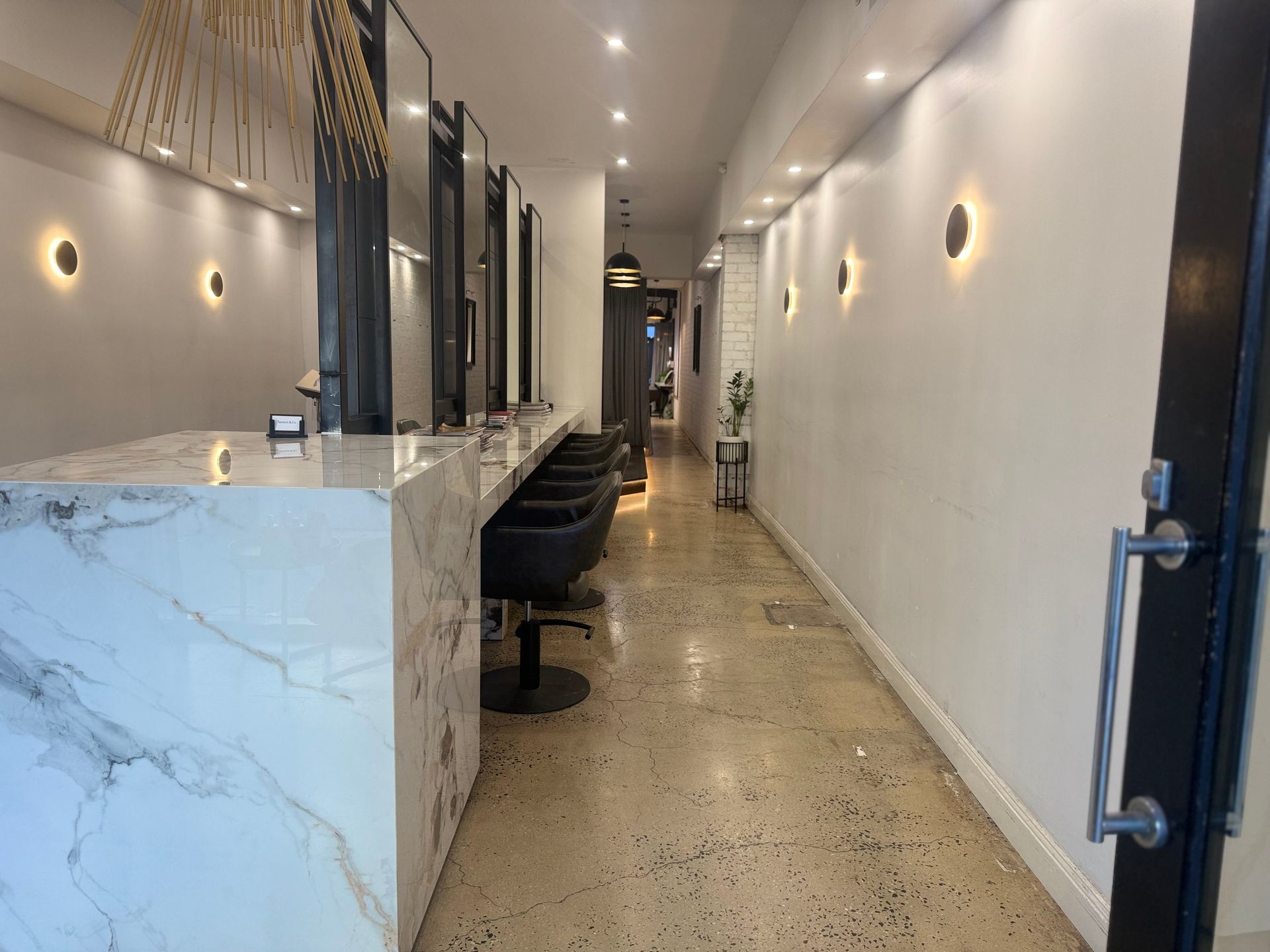 Narrow modern hallway with marble counter on left, bar stools, wall sconces, and polished concrete floor.
