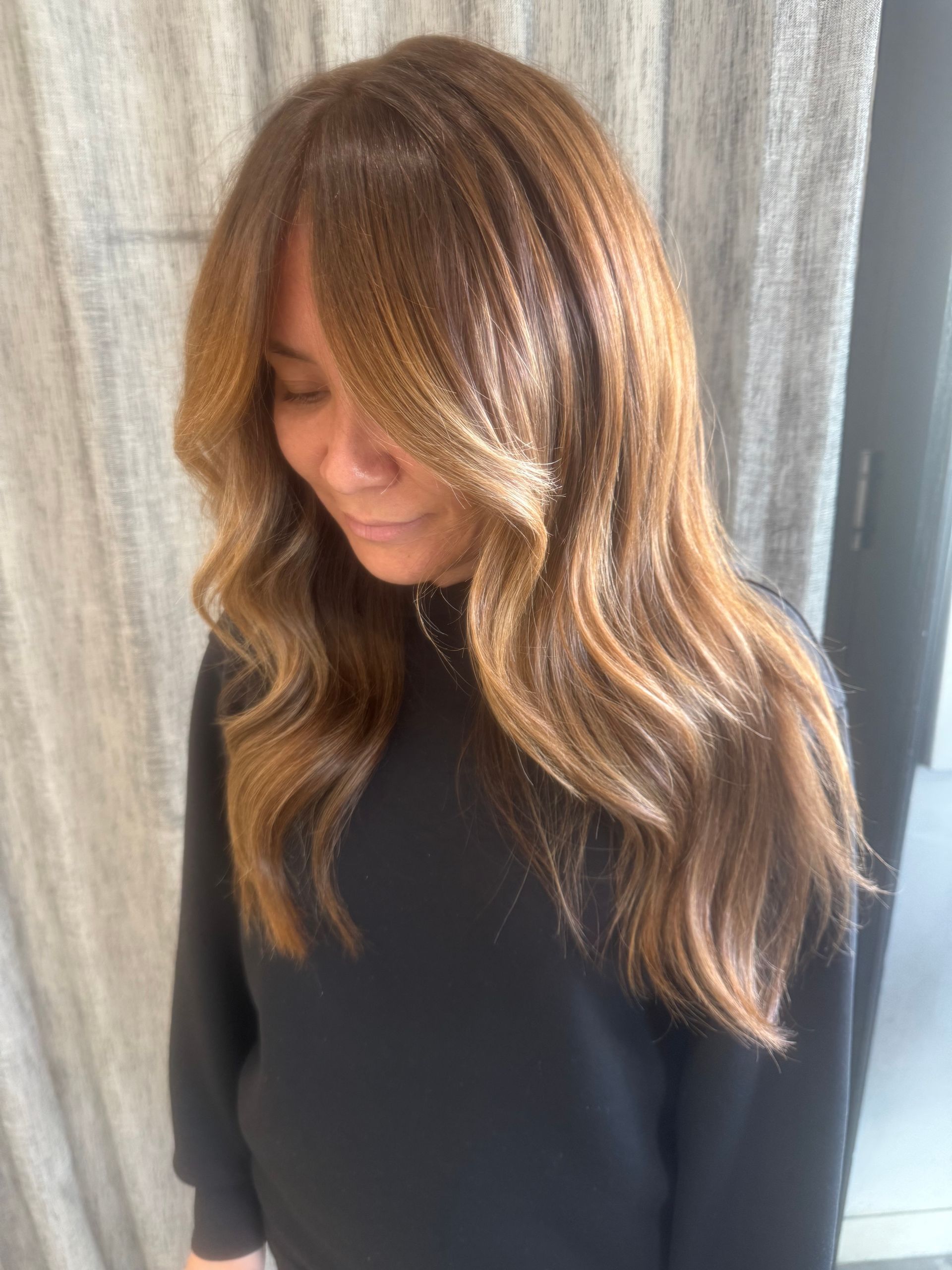 Woman with long wavy light-brown hair looking down, wearing a black top against a gray curtain.