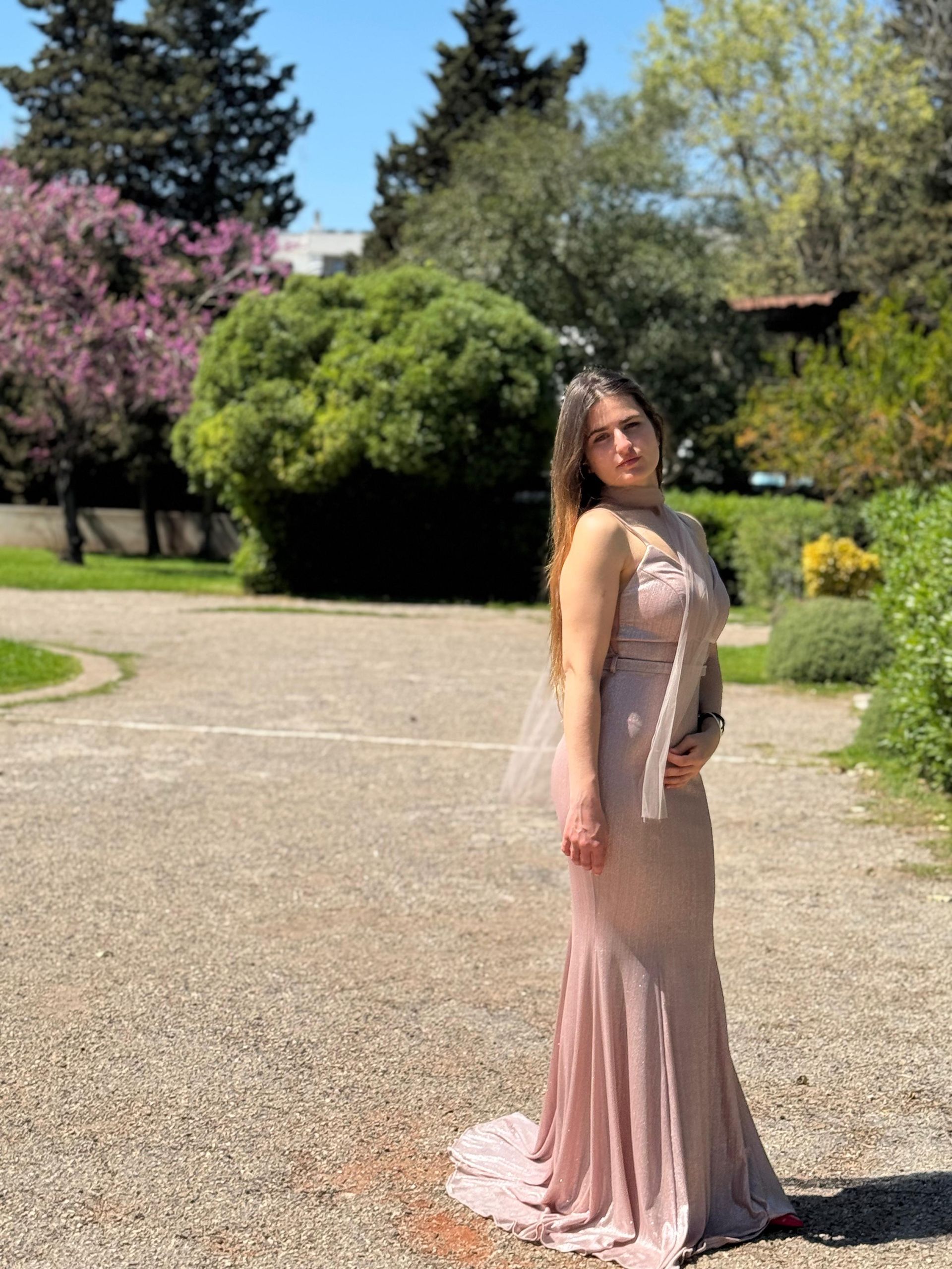 Una donna con un lungo abito color cipria è in piedi su un sentiero di ghiaia illuminato dal sole in un giardino.