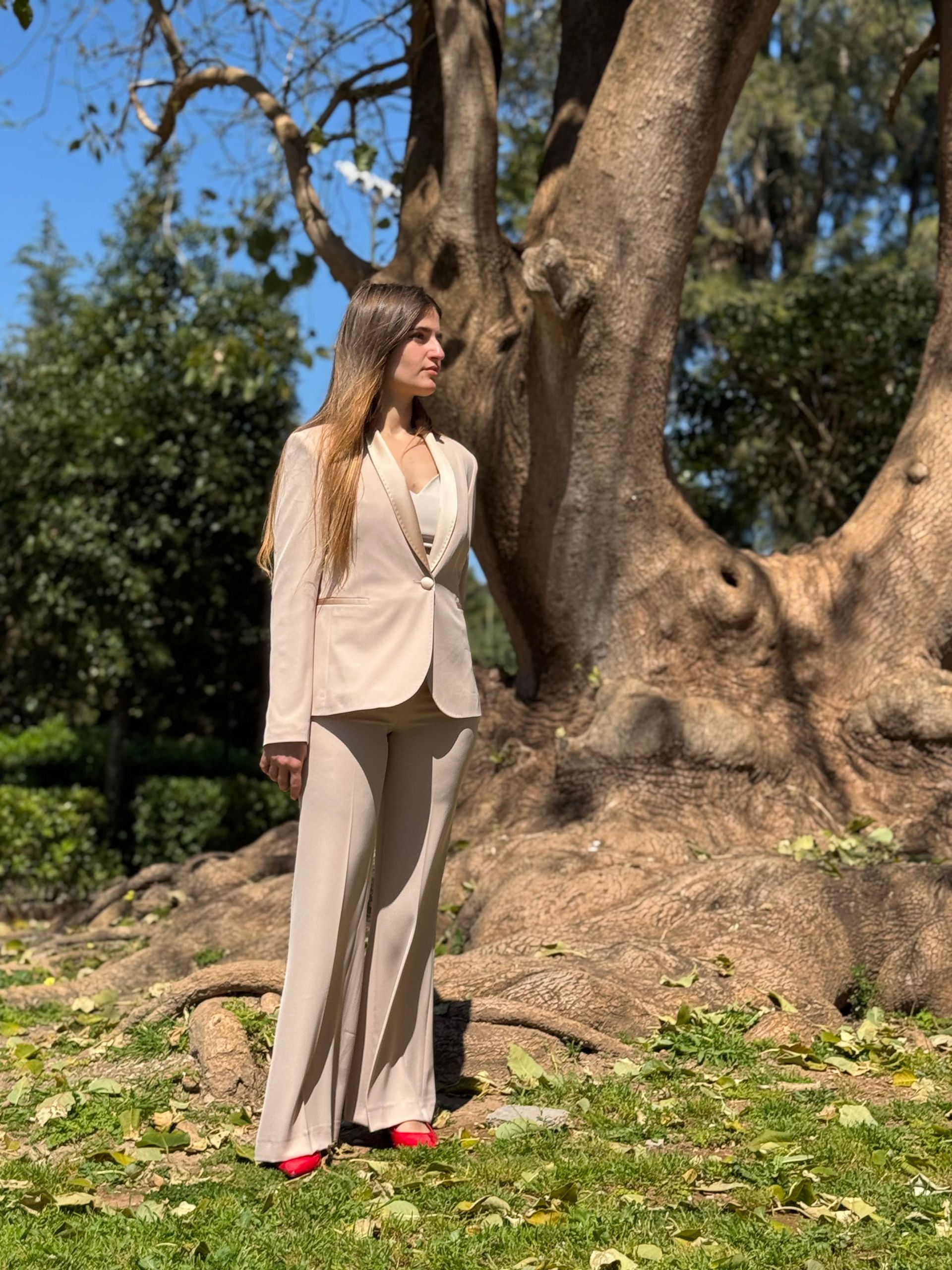 Una donna in tailleur beige è in piedi accanto a un grande albero in un parco soleggiato, con indosso scarpe rosse.