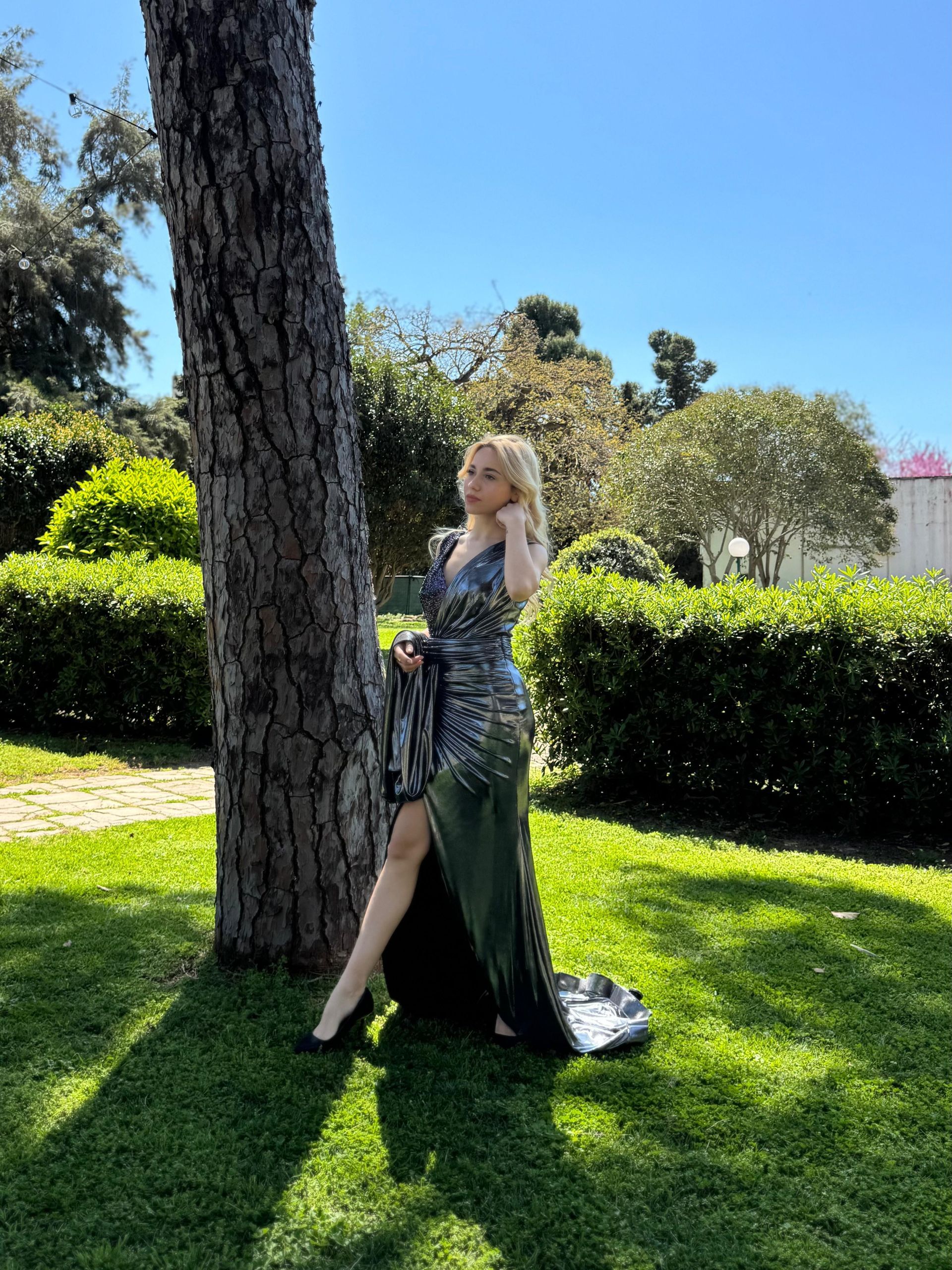 Una donna con indosso un abito verde scuro è appoggiata a un albero in un parco soleggiato, con siepi e prato alle sue spalle.