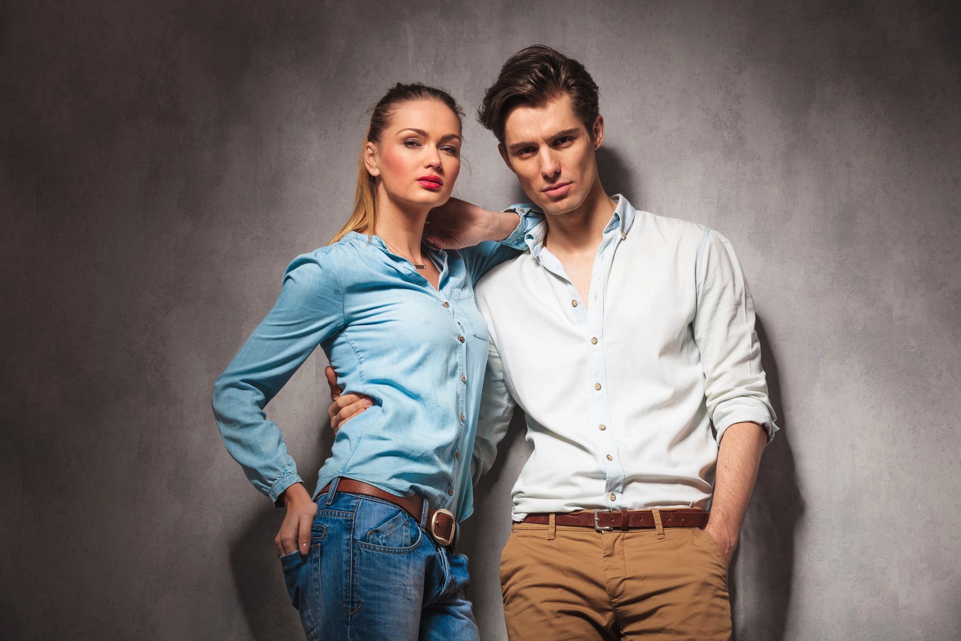 Due persone vestite in modo informale posano davanti a uno sfondo grigio in studio: una con una camicia blu e jeans, l'altra con una camicia bianca e pantaloni beige.