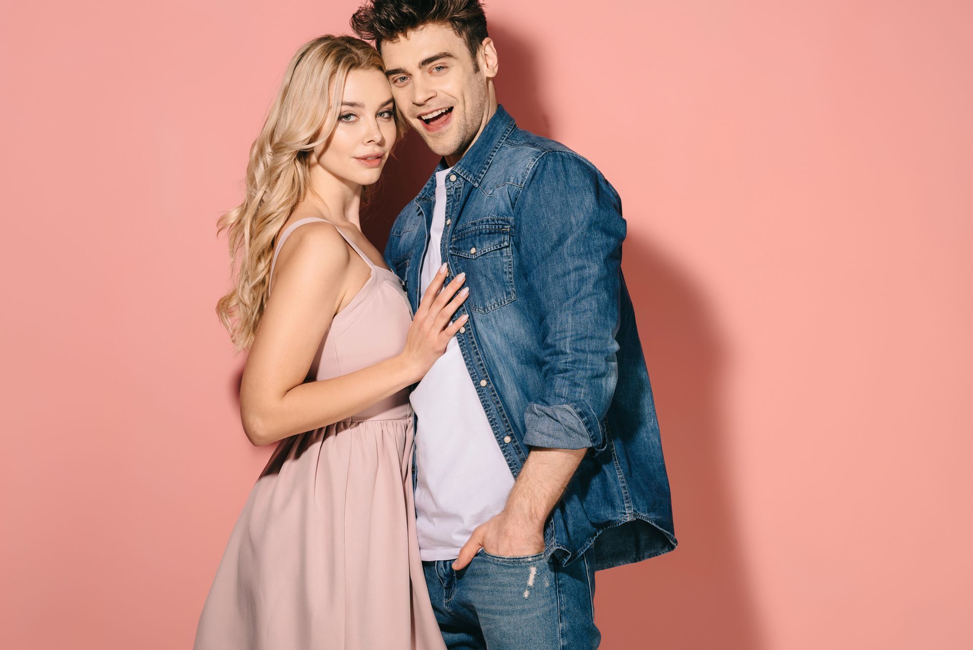Una coppia sorridente posa insieme su uno sfondo rosa, la donna con un abito color cipria e l'uomo con una giacca di jeans.