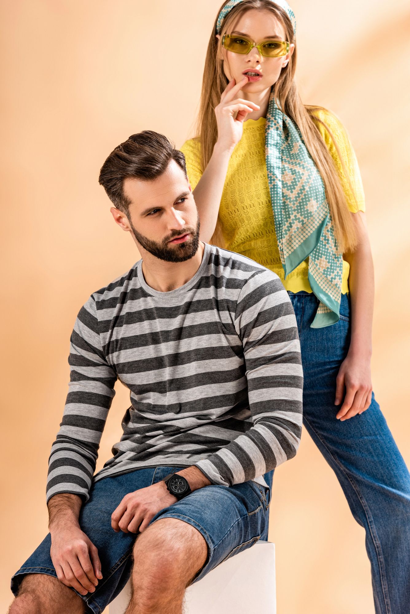 Due modelle in posa davanti a uno sfondo color pesca, una seduta con indosso un abito a righe, l'altra in piedi con un abito giallo e blu.