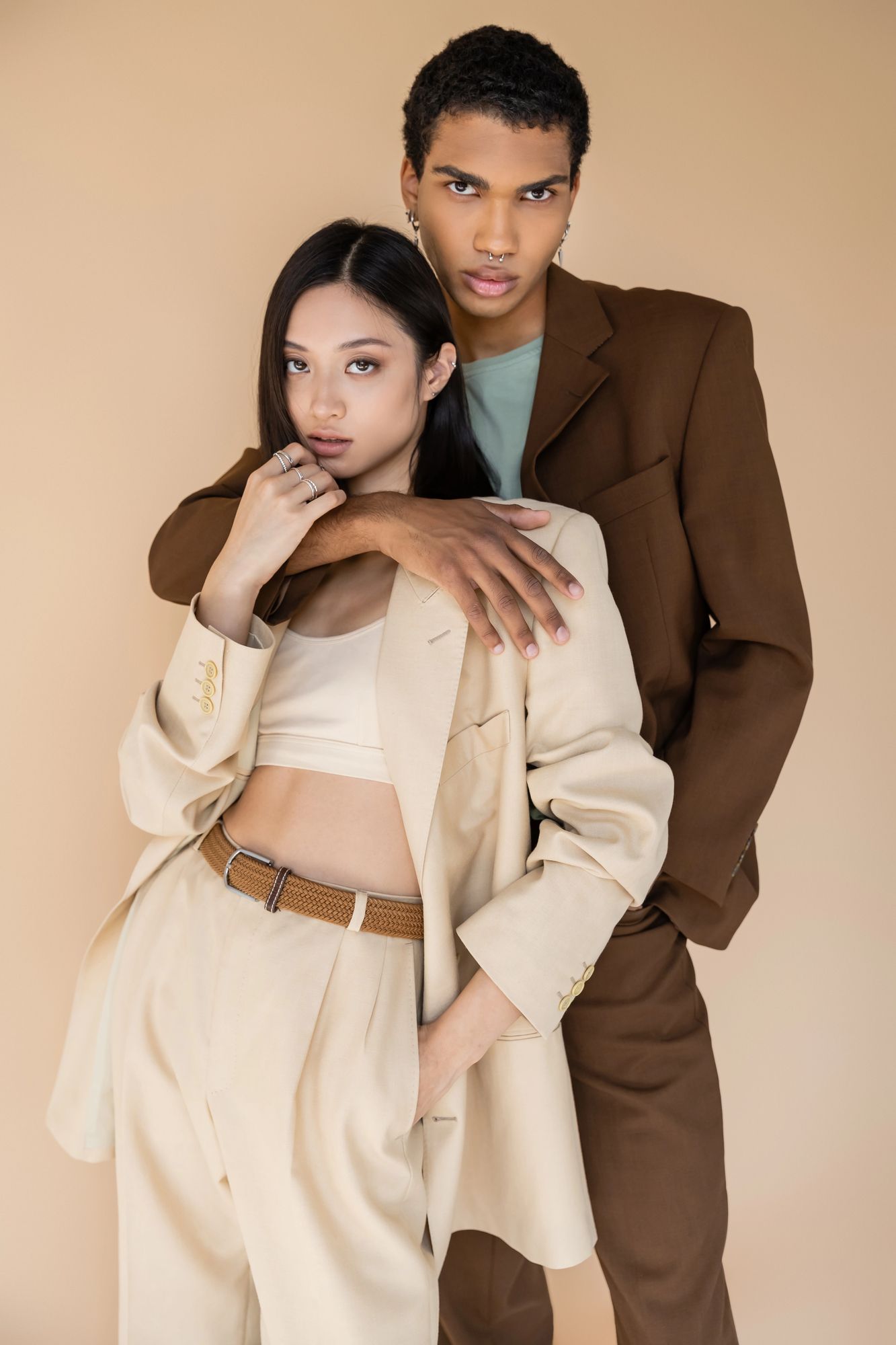 Due modelle, vestite con abiti sartoriali beige e marroni, posano davanti a uno sfondo color sabbia in studio.