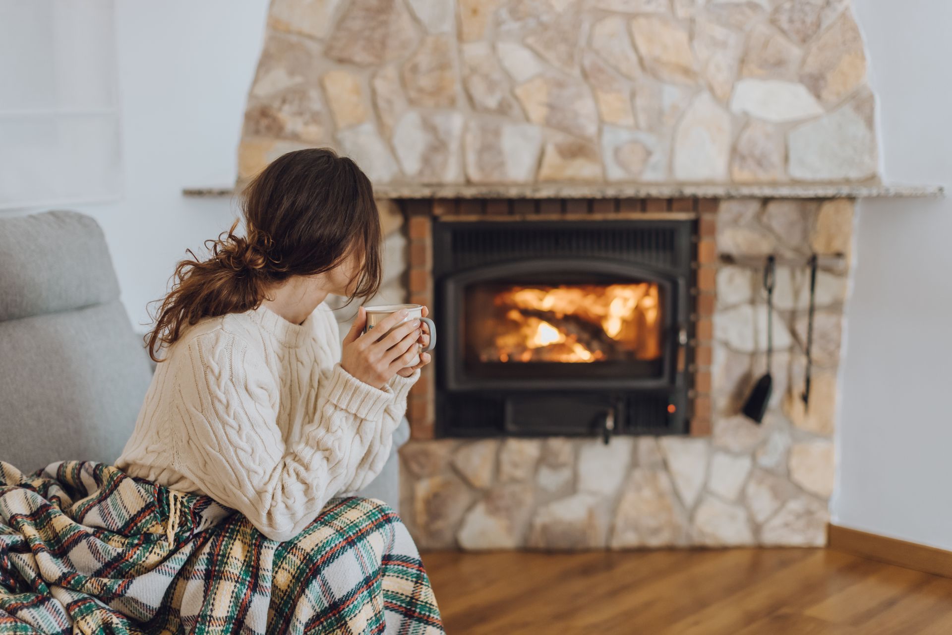 A woman sitting at home by a gas fireplace with a hot beverage and warming her hands.