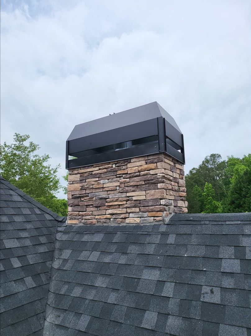 Chimney with dark metal cap on a stone facade, rising above a gray shingled roof, set against a cloudy sky.