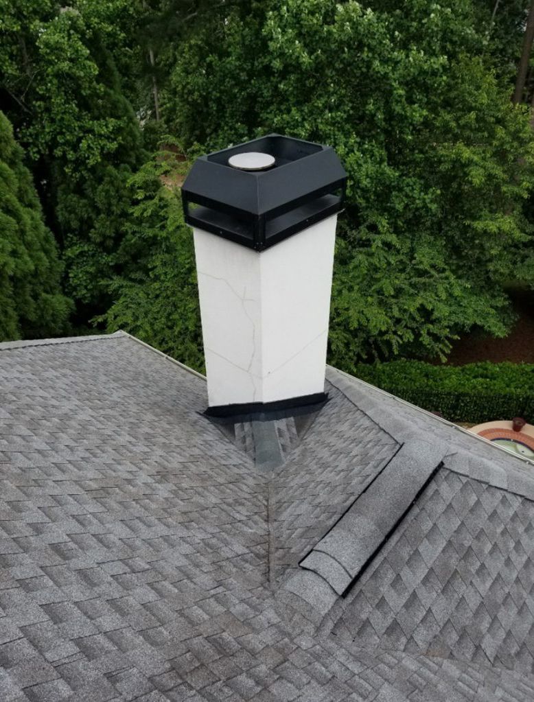 White chimney with black cap on a gray shingled roof, trees in the background.