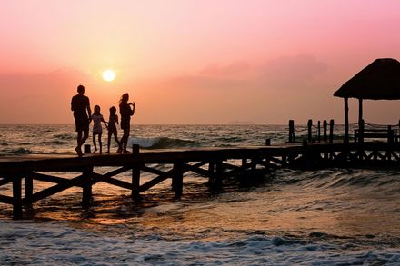 Silhouetted people on a pier at sunset over the sea, with a small hut on the right.