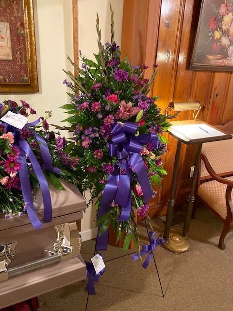 A floral funeral standing spray with purple and pink flowers and a large purple bow, displayed on a stand in a room.