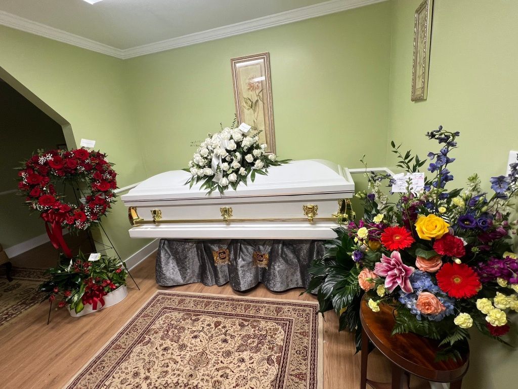 A white casket with a white floral arrangement sits on a stand in a green-walled room, flanked by large floral displays.
