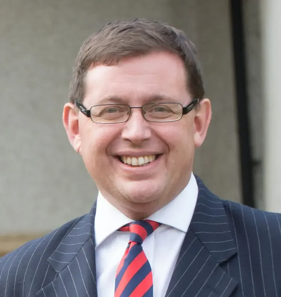 Smiling man in glasses and a suit with a red-and-blue striped tie outdoors