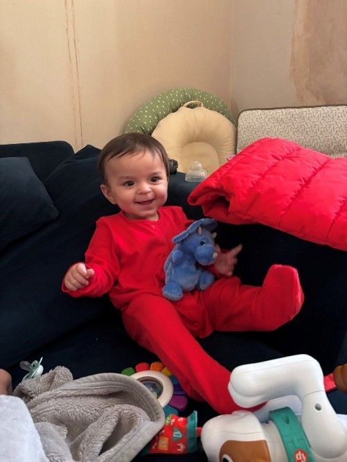 Smiling baby in red pajamas sitting on a couch, holding a blue stuffed toy, with pillows and toys nearby