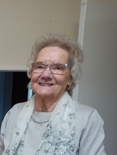 Smiling woman wearing glasses and a light scarf, standing indoors near a doorway.