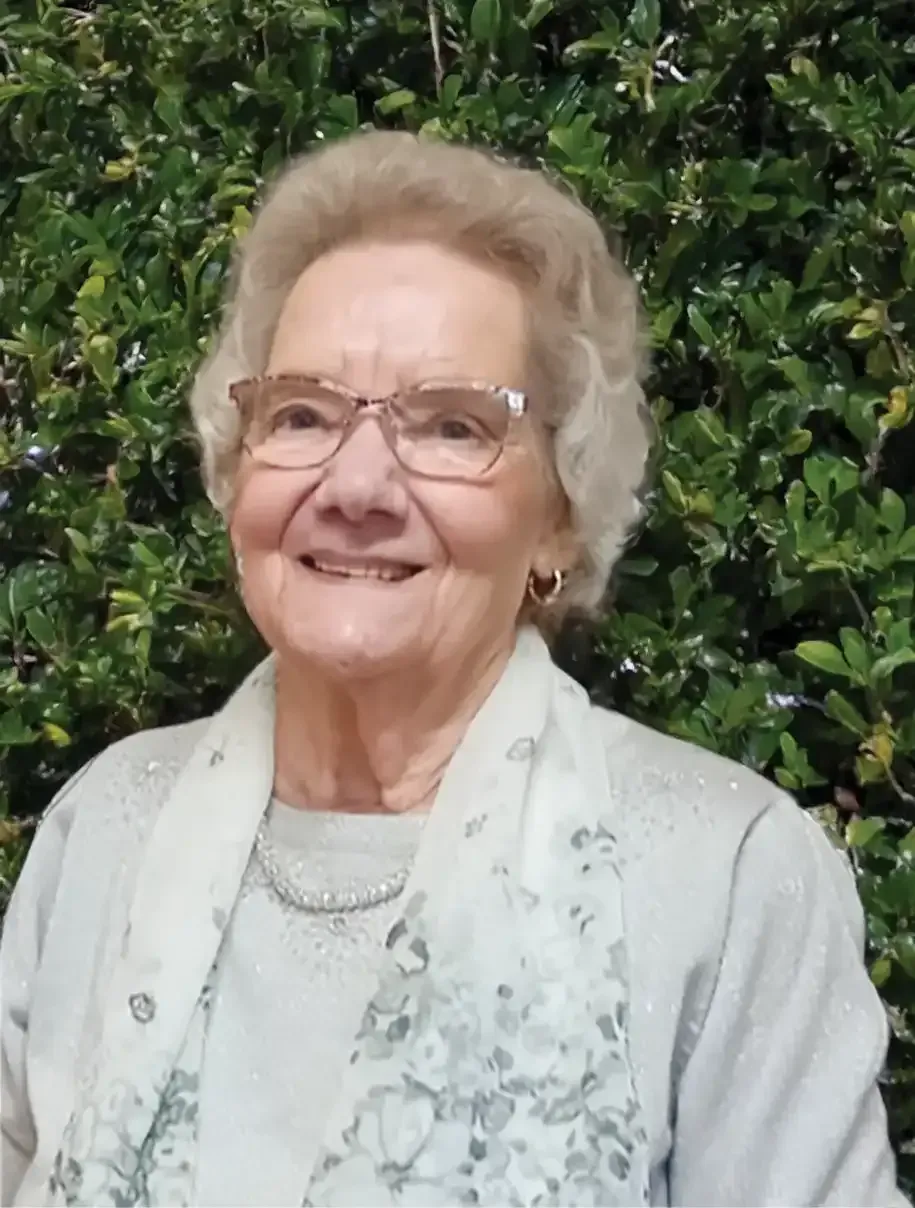 Smiling woman in glasses and a light scarf standing before green foliage.